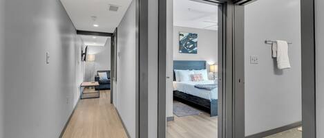 A modern hotel corridor with a wooden floor, a bed with a blue headboard, and a painting on the wall.