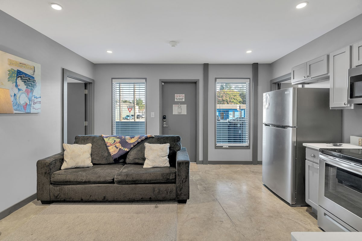 A modern kitchen with grey cabinets, a stainless steel refrigerator, and an oven. There is a grey sofa with patterned cushions in the living area.
