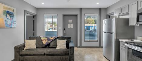 A modern kitchen with grey cabinets, a stainless steel refrigerator, and an oven. There is a grey sofa with patterned cushions in the living area.