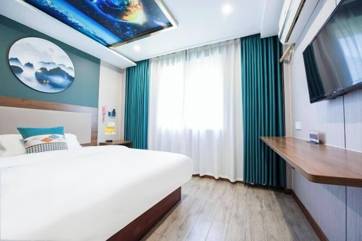 Una habitación de hotel con cama, televisión, un cuadro y ventana con cortinas.
