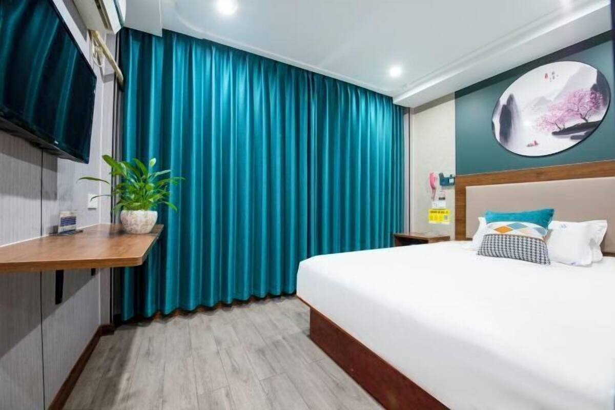 Habitación de hotel con cama, cortinas color turquesa, televisor montado en la pared, planta en maceta y una obra de arte en la pared.