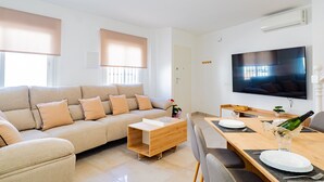 A modern living room with a sofa, a wooden coffee table, a dining table set for two, and a flat-screen TV mounted on the wall. - Cubo’s La Perla Town Center House / urban / air conditioning / free Wi-Fi (Alhaurín el Grande)