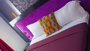 Ein Hotelzimmer mit violettem Kopfteil, einem Bett mit burgunderfarbener Bettdecke und zwei dekorativen Kissen mit bunten Mustern.