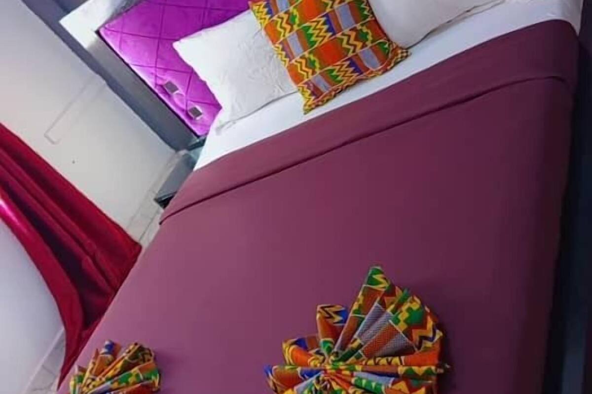 Une chambre d’hôtel avec une tête de lit violette, un lit recouvert d’une courtepointe bordeaux et deux coussins décoratifs aux motifs colorés.