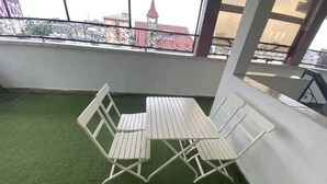 Ein Balkon mit einem weißen Metalltisch und -stühlen auf Kunstgrün, mit Blick auf eine Stadtlandschaft mit Gebäuden und einem Turm.