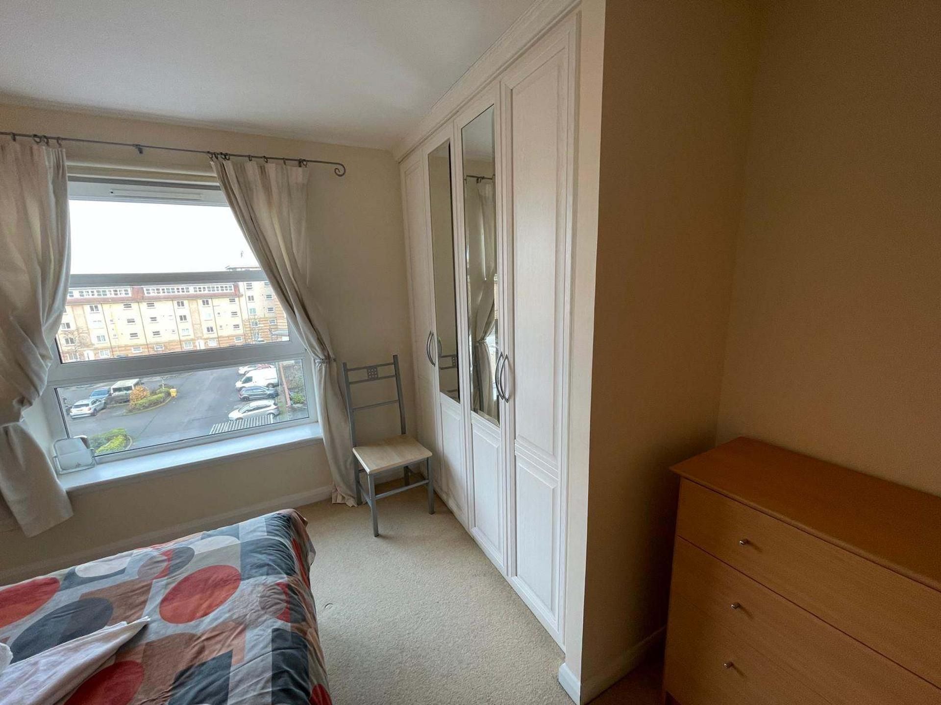 Ein Schlafzimmer mit Bett, Kommode, einem Fenster mit Vorhängen und Blick auf die Straße.