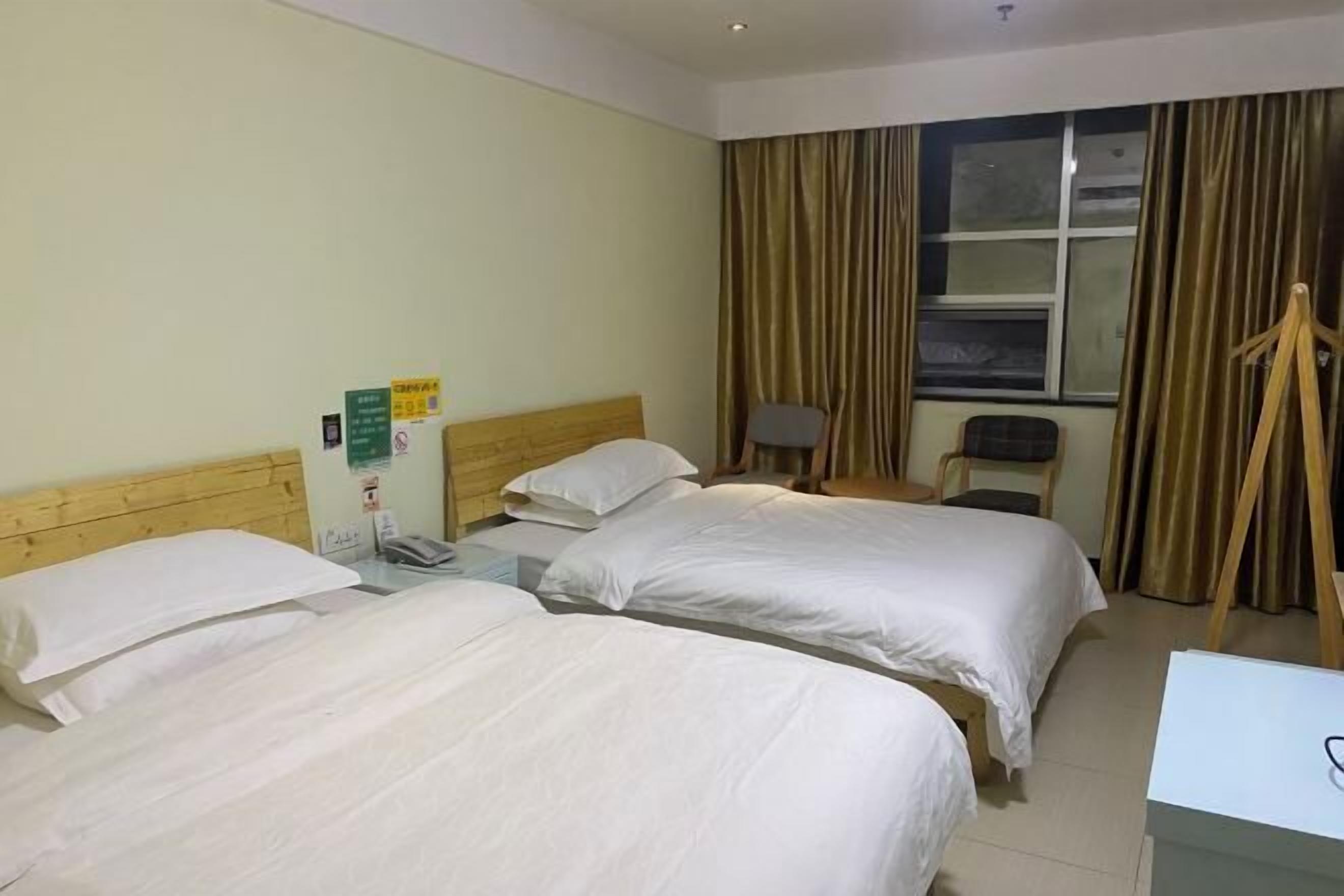 Quarto de hotel com duas camas de solteiro, cabeceira de madeira, escrivaninha com cadeira, janela com cortinas e uma mesinha de cabeceira com abajur.