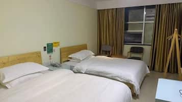 A hotel room with two single beds, a wooden headboard, a desk with a chair, a window with curtains, and a small table with a lamp.