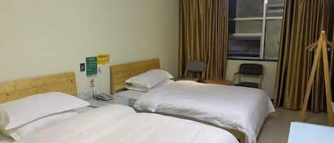 Quarto de hotel com duas camas de solteiro, cabeceira de madeira, escrivaninha com cadeira, janela com cortinas e uma mesinha de cabeceira com abajur.