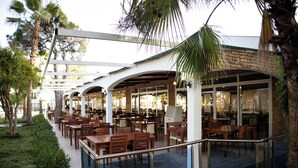 An outdoor dining area with multiple wooden tables and chairs, surrounded by palm trees and a covered walkway. - Club Golf Hotel - All Inclusive (Manavgat)