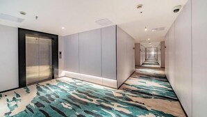 Moderni hotellikäytävä, jossa on kuvioitu matto, keskeisellä paikalla sijaitseva hissi ja pitkä käytävä, jonka molemmilla puolilla on ovia.