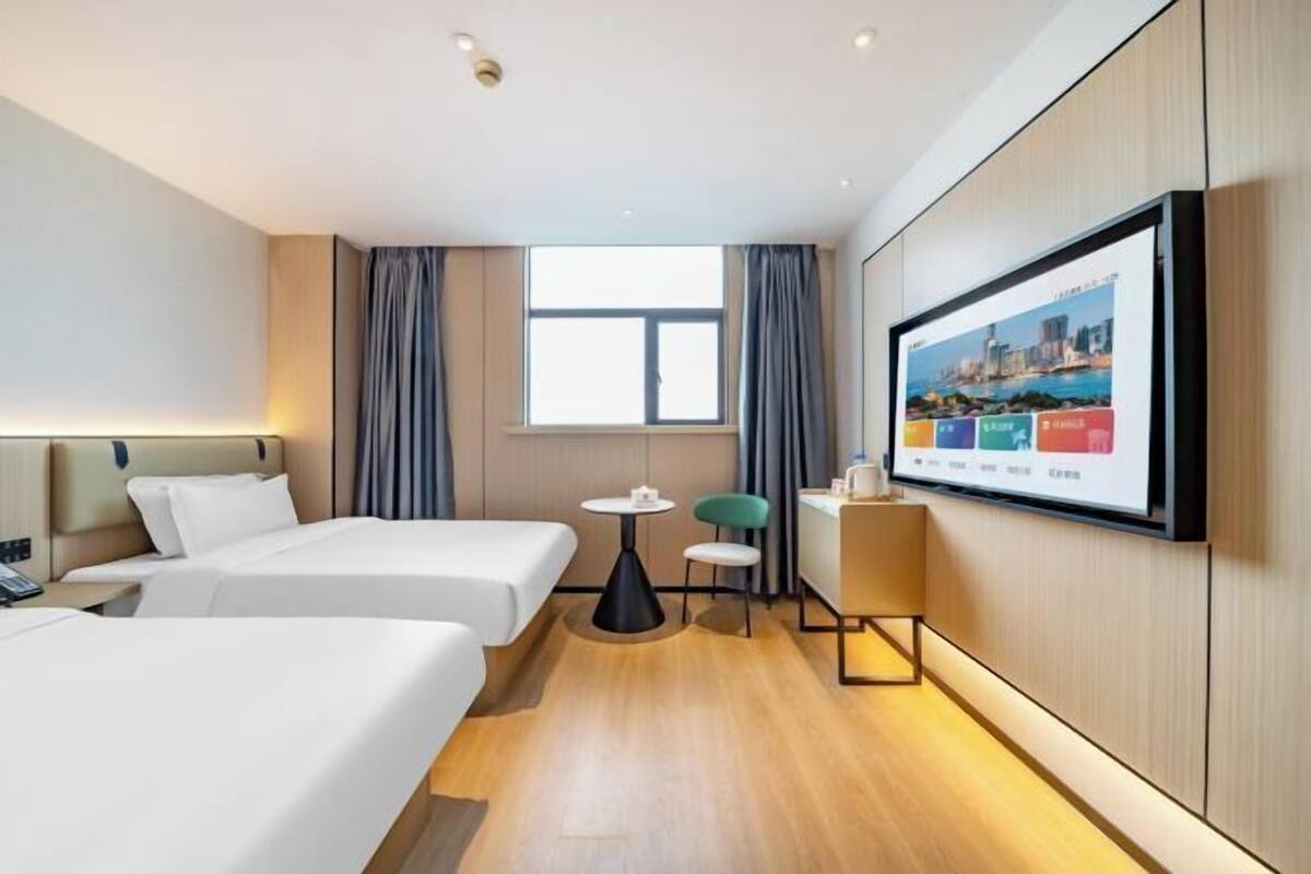 Een hotelkamer met twee bedden, een klein tafeltje, een stoel, een aan de muur bevestigde televisie en een raam met gordijnen.