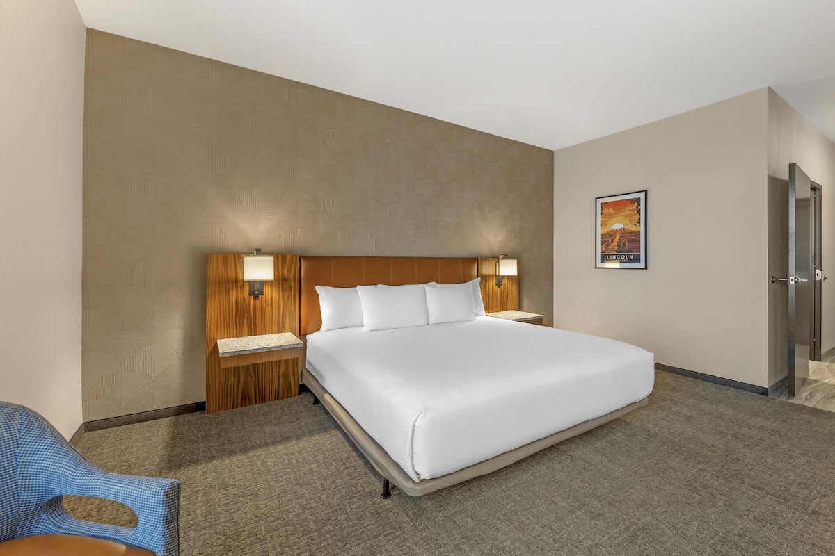 A hotel room with a large bed, wooden headboard, two bedside lamps, a framed picture on the wall, and a door to the right.