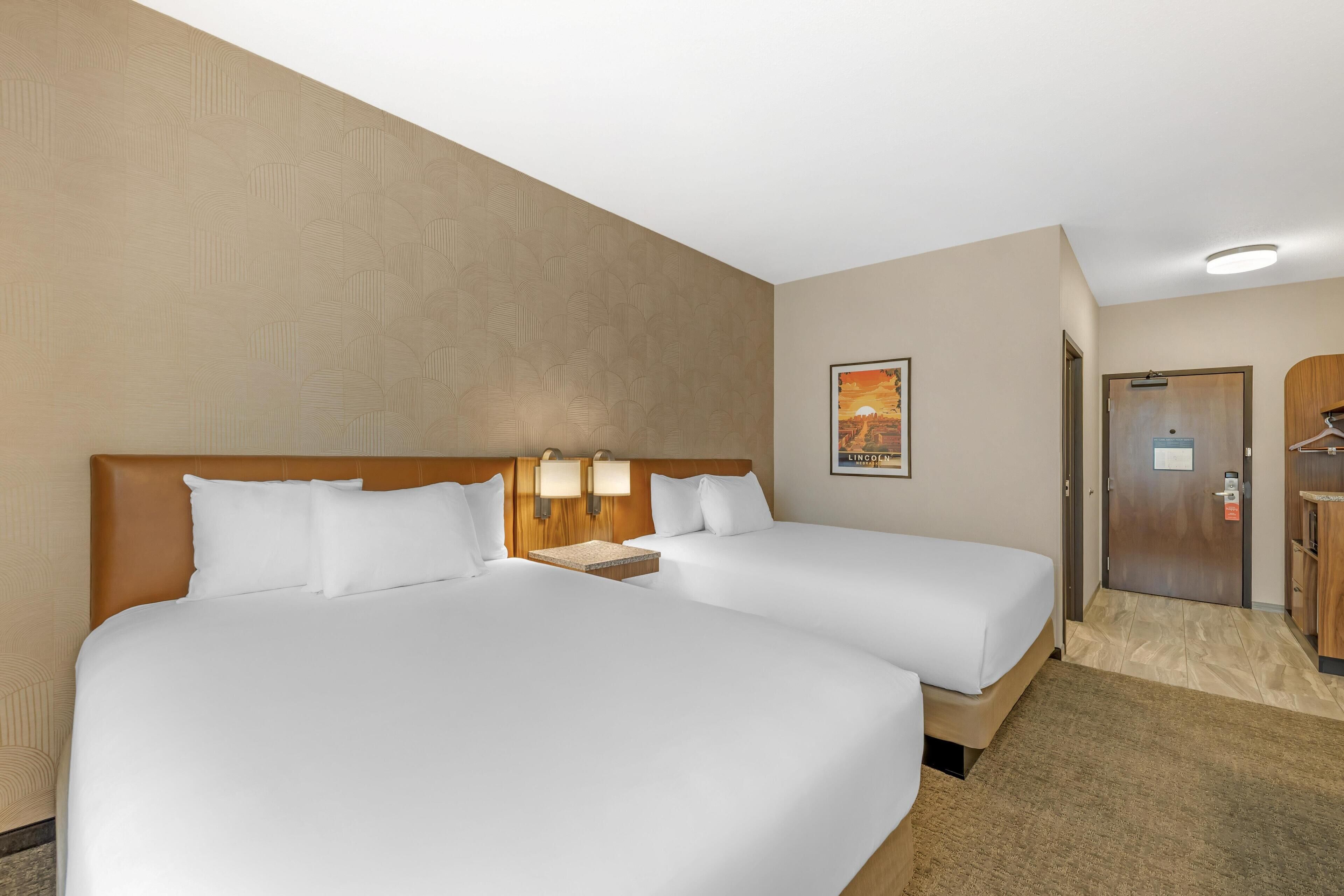 A hotel room with two beds, a wooden headboard, a nightstand with lamps, a door with a coat rack, and a poster on the wall.