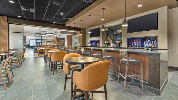 A modern restaurant with multiple tables and chairs, a bar area with a counter and shelves of bottles, and a large screen on the wall.