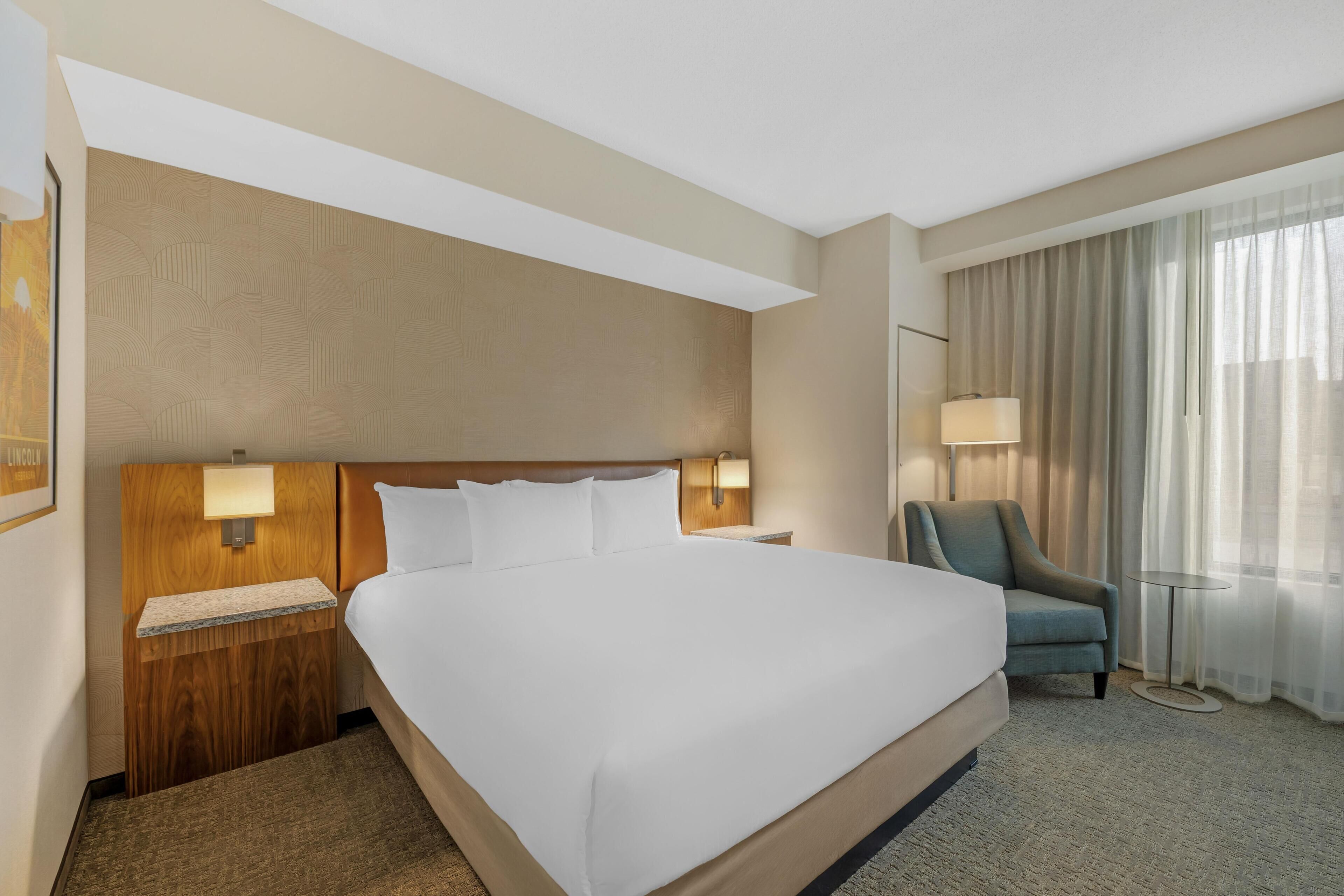 Een hotelkamer met een groot bed, een houten hoofdbord, een nachtkastje met een lamp, een stoel en een raam met gordijnen.