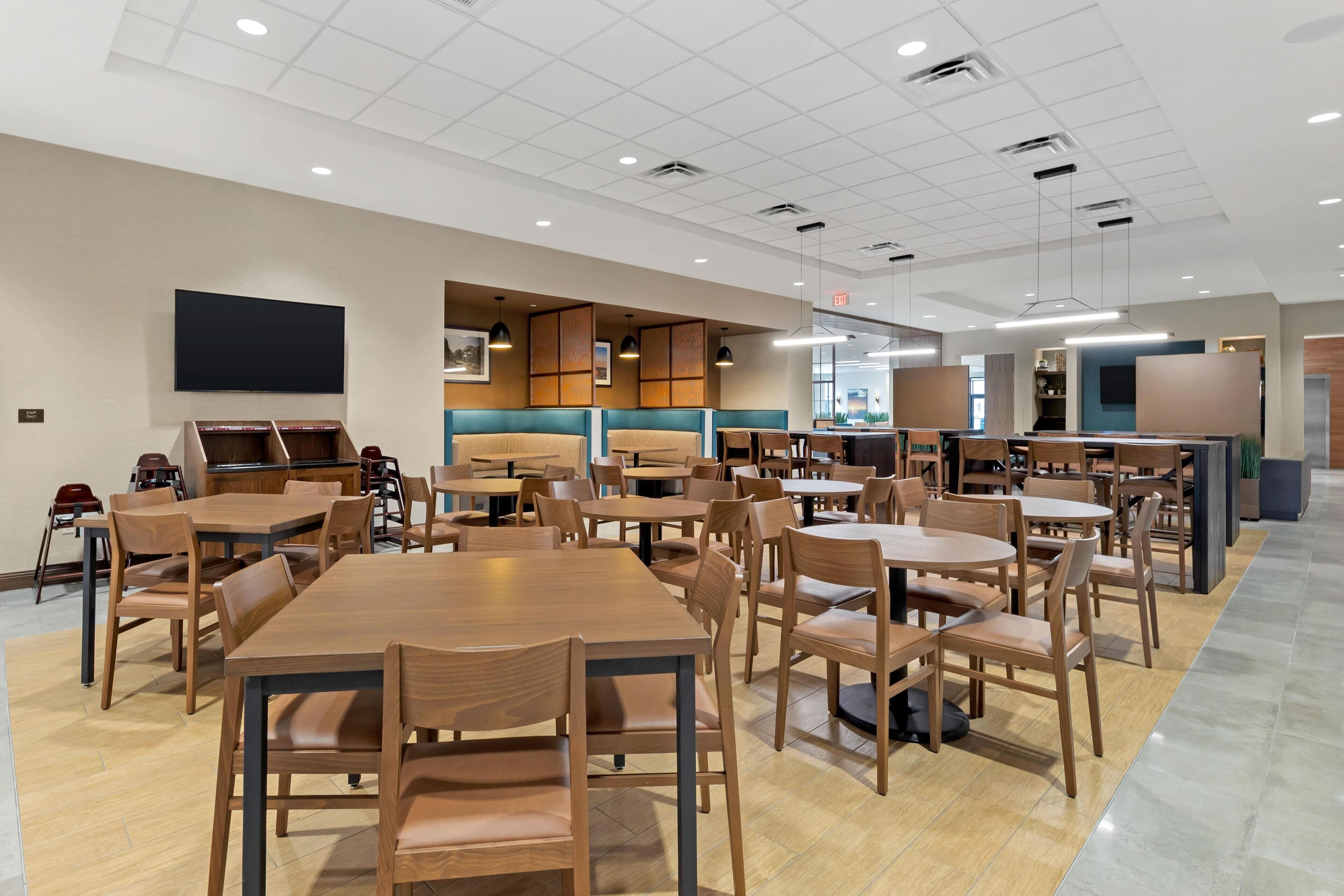 A spacious dining area with multiple wooden tables and chairs, a flat-screen TV mounted on the wall, and a modern ceiling design with recessed lighting.
