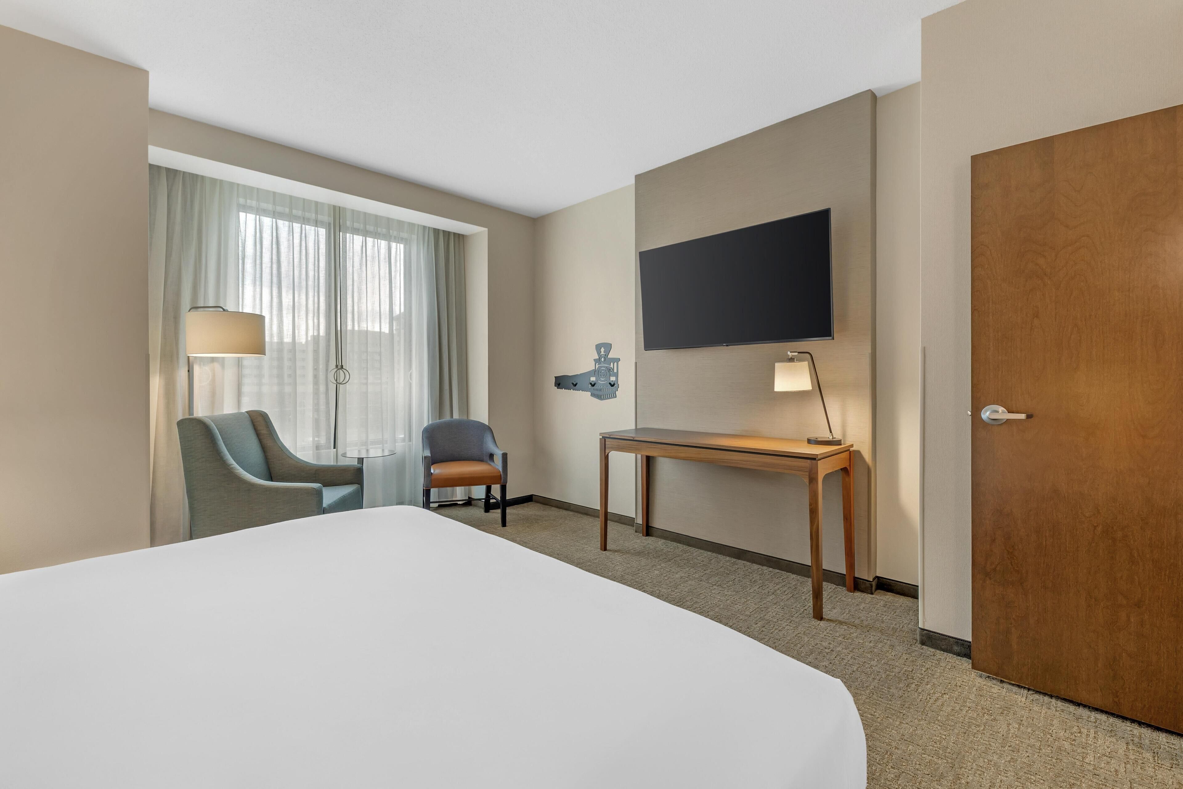 A hotel room with a large bed, a desk, a chair, a TV mounted on the wall, and a door.