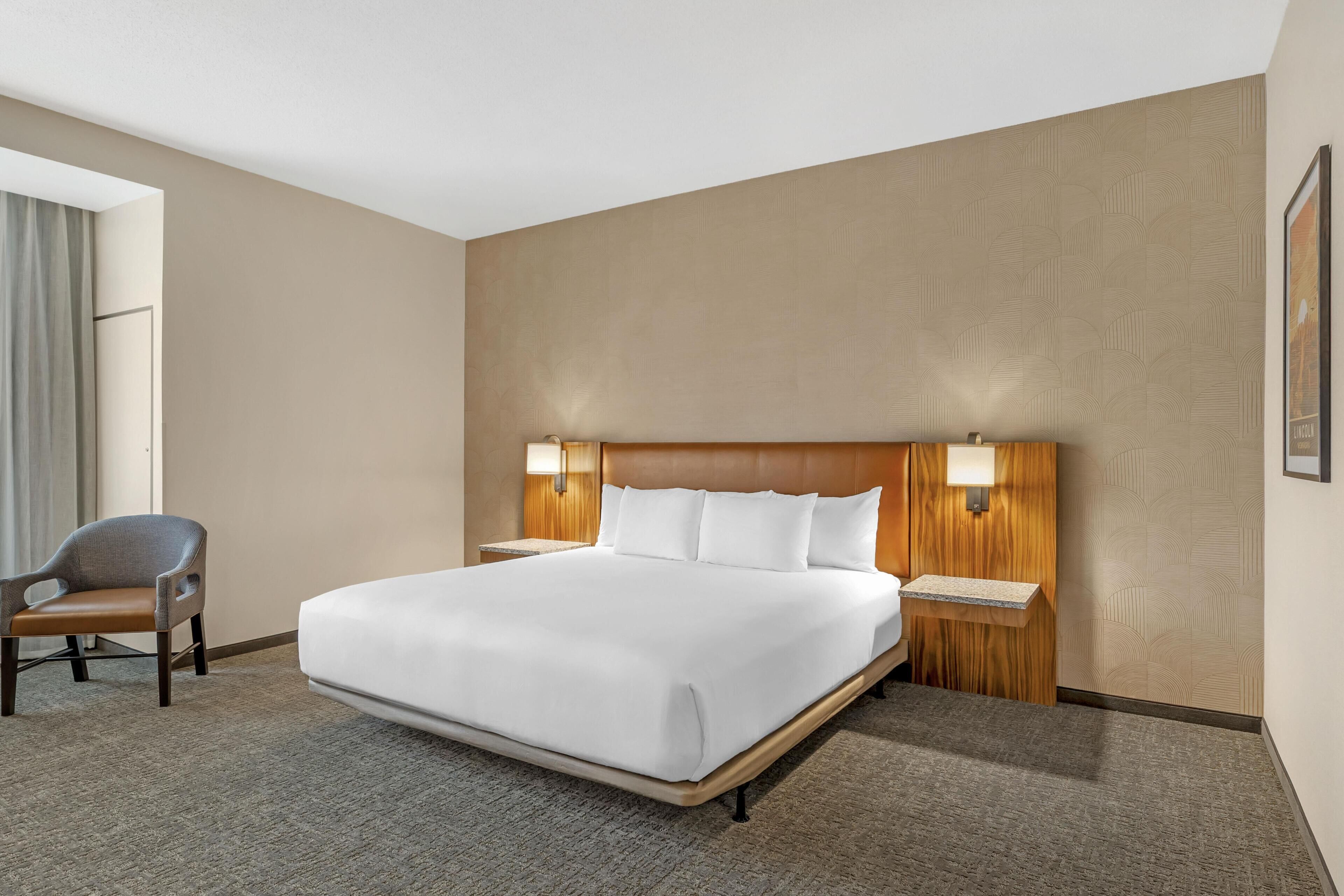 A hotel room with a large bed, two bedside tables, a chair, and a window with curtains.