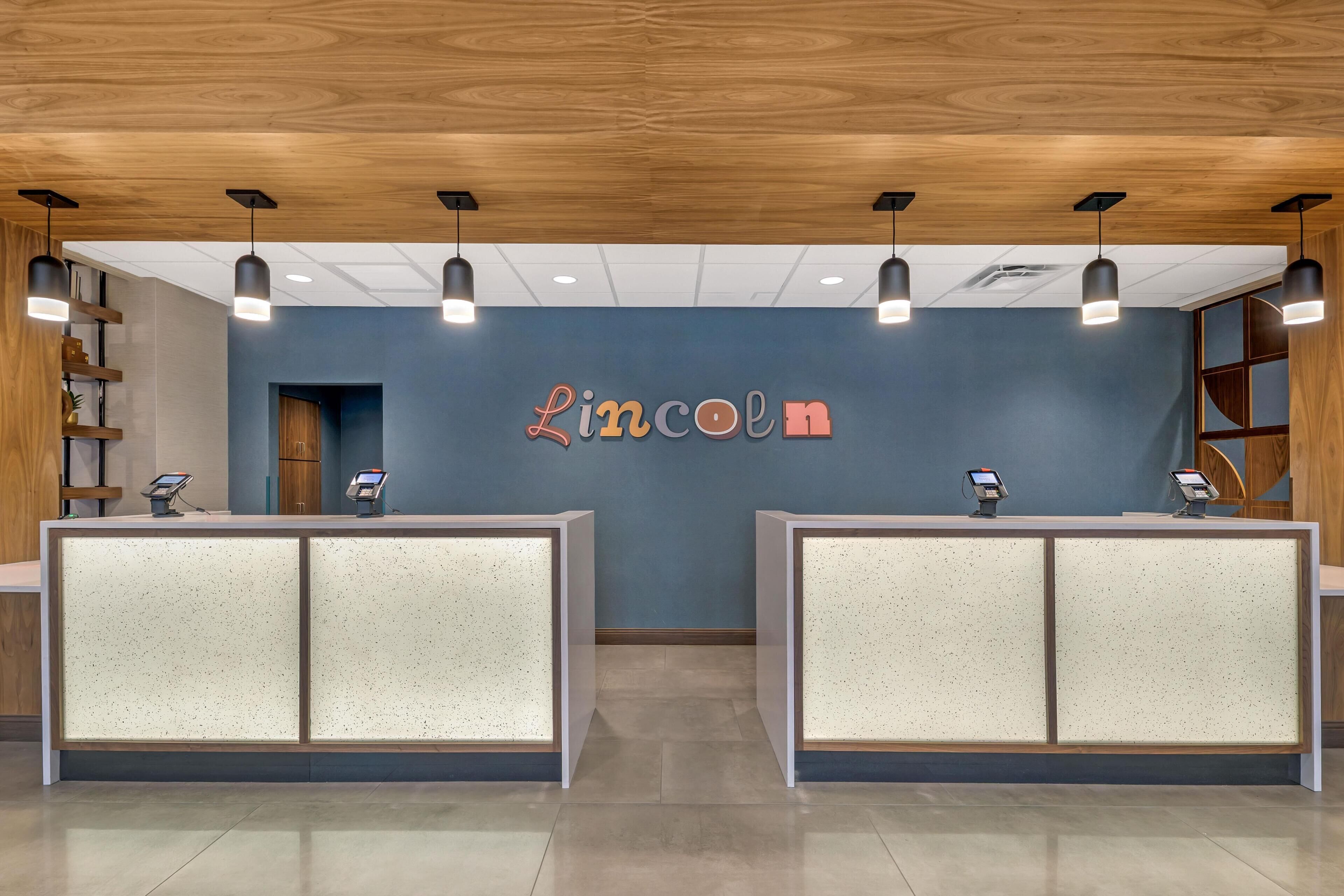 Un'area reception con soffitto in legno, due scrivanie per la reception con chioschi digitali e una parete blu con la scritta "Lincoln" al centro.