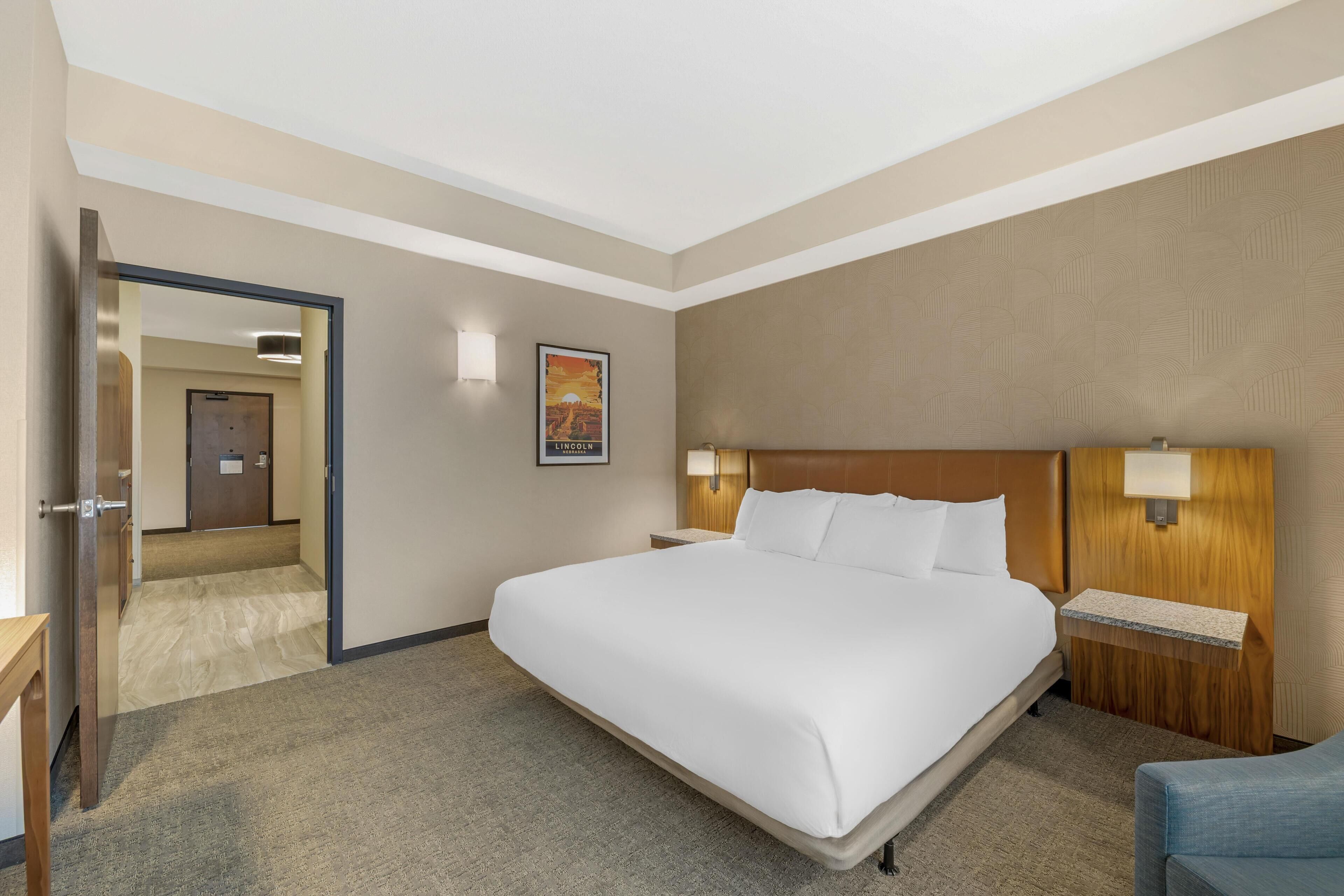 A hotel room with a large bed, a wooden headboard, a nightstand with a lamp, and a view of the hallway.