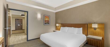 A hotel room with a large bed, a wooden headboard, a nightstand with a lamp, and a view of the hallway.