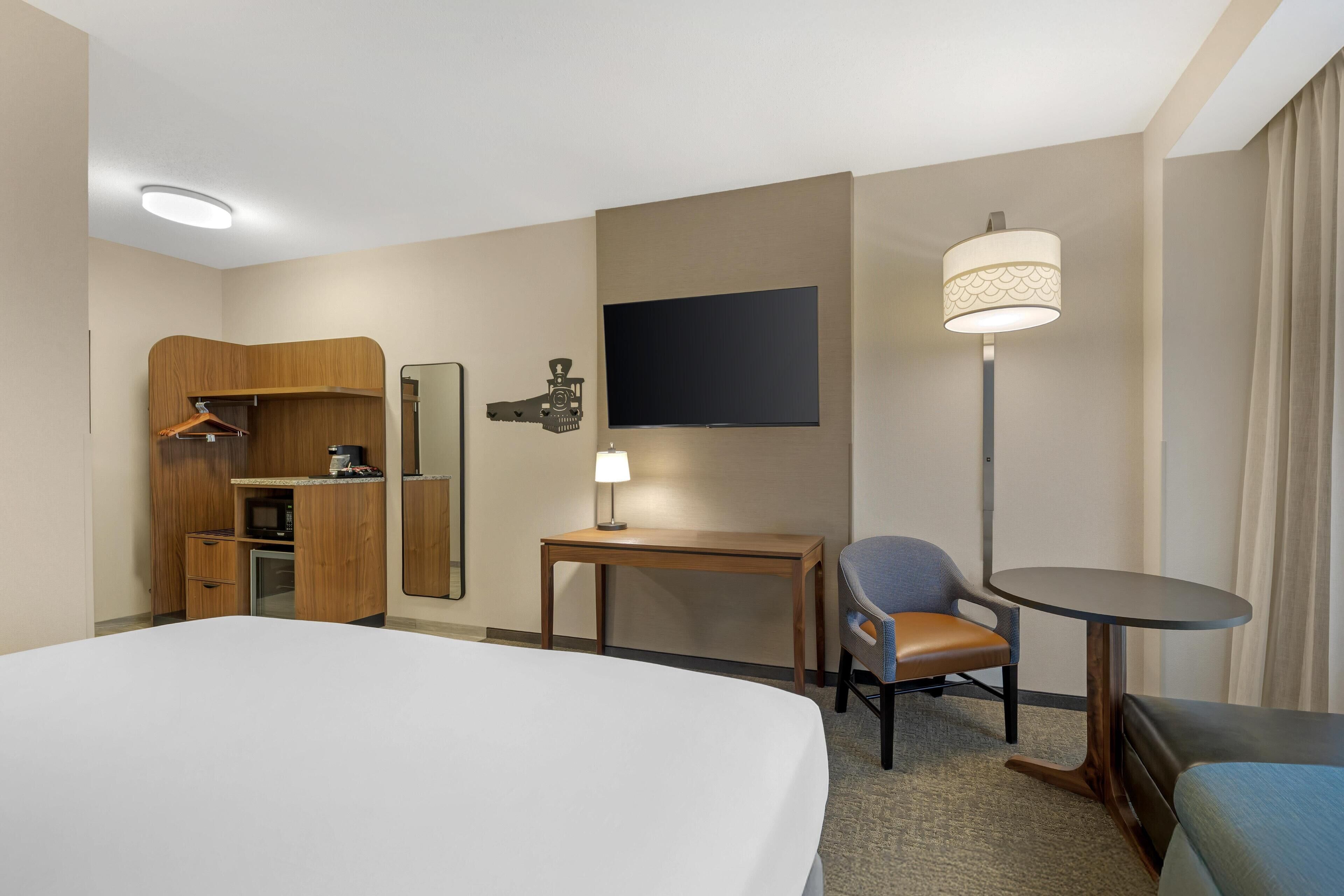 A hotel room with a large bed, a desk with a chair, a small table with a lamp, a wardrobe, and a TV mounted on the wall.