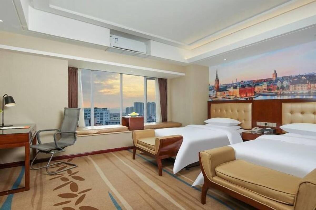 A hotel room with two beds, a desk, a chair, a window with a cityscape view, and an air conditioning unit.