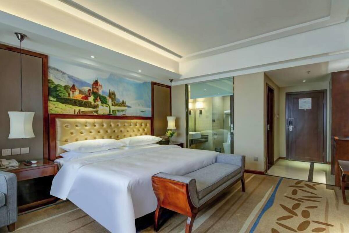 A hotel room with a large bed, a desk with a lamp, a painting on the wall, and a view of the bathroom through a glass door.