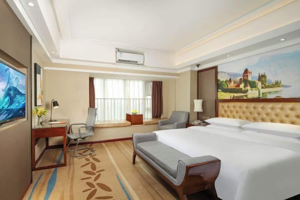 A hotel room with a large bed, a desk with a chair, a TV, and a painting of a castle on the wall.