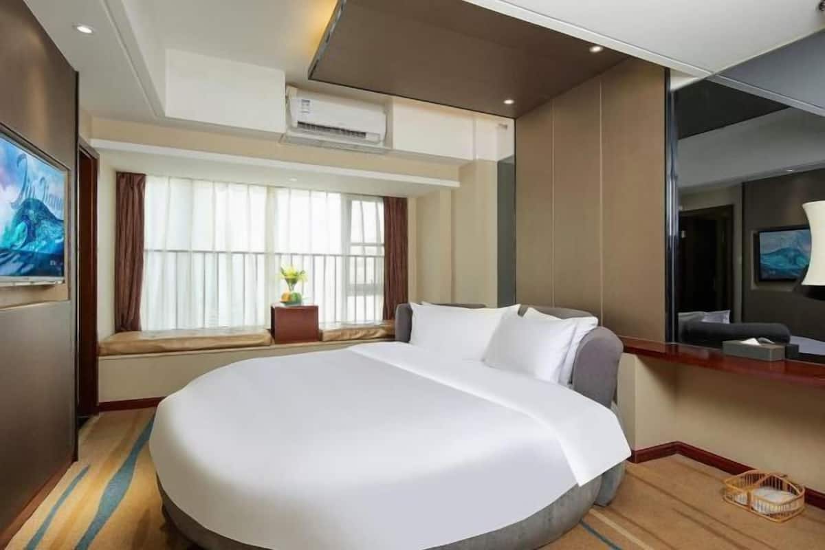 A hotel room with a large bed, a window with curtains, an air conditioning unit, a television, and a small table with a basket.