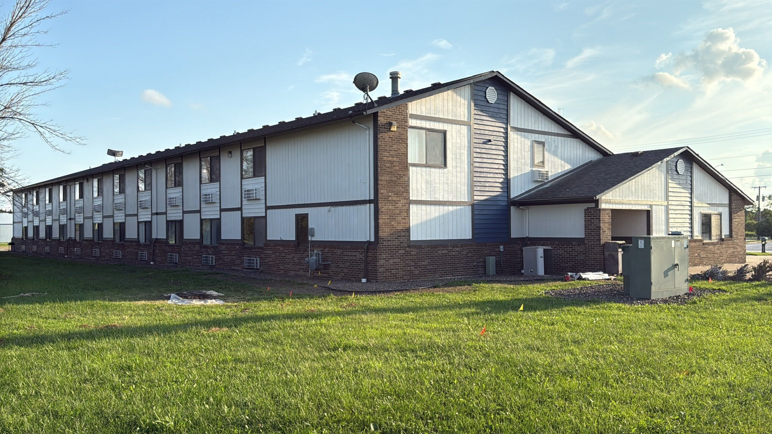 A long, single-story building with a brick foundation and white siding, featuring multiple windows and a satellite dish on the roof.