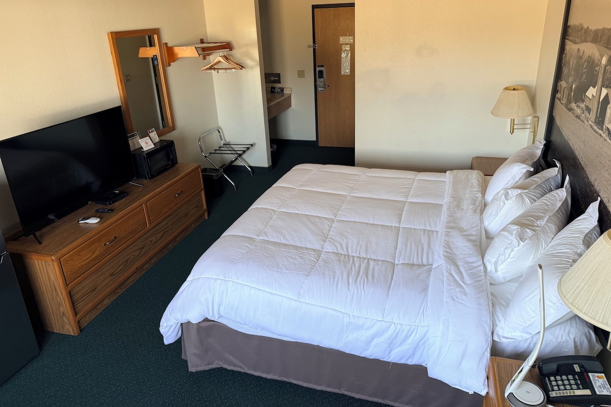 Et hotelværelse med en stor seng, en trækommode, et fjernsyn, et skrivebord med lampe og et billede på væggen.