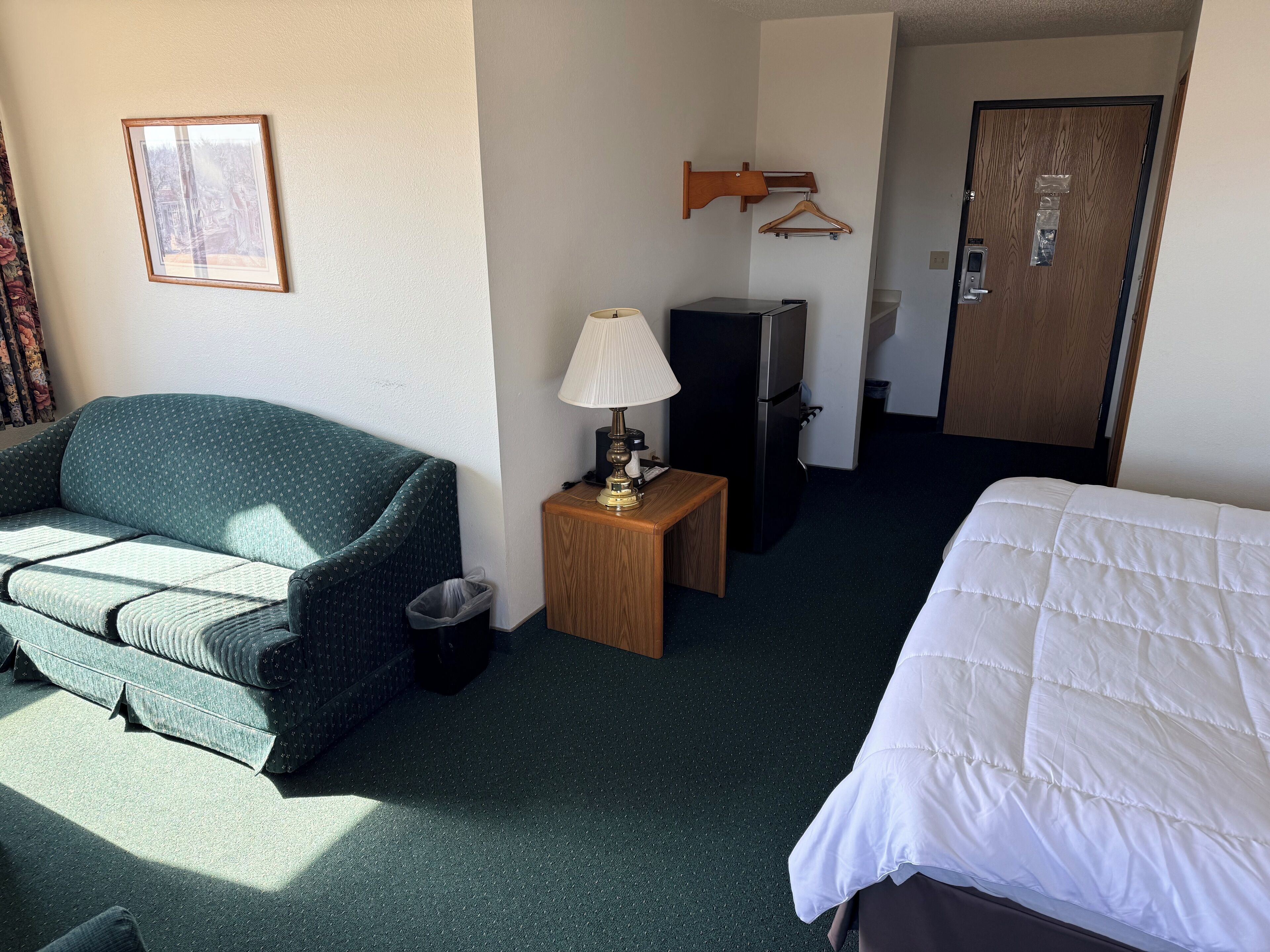 Habitación de hotel con un sofá verde, una cama blanca, una mesita de madera con una lámpara, un televisor y un perchero.
