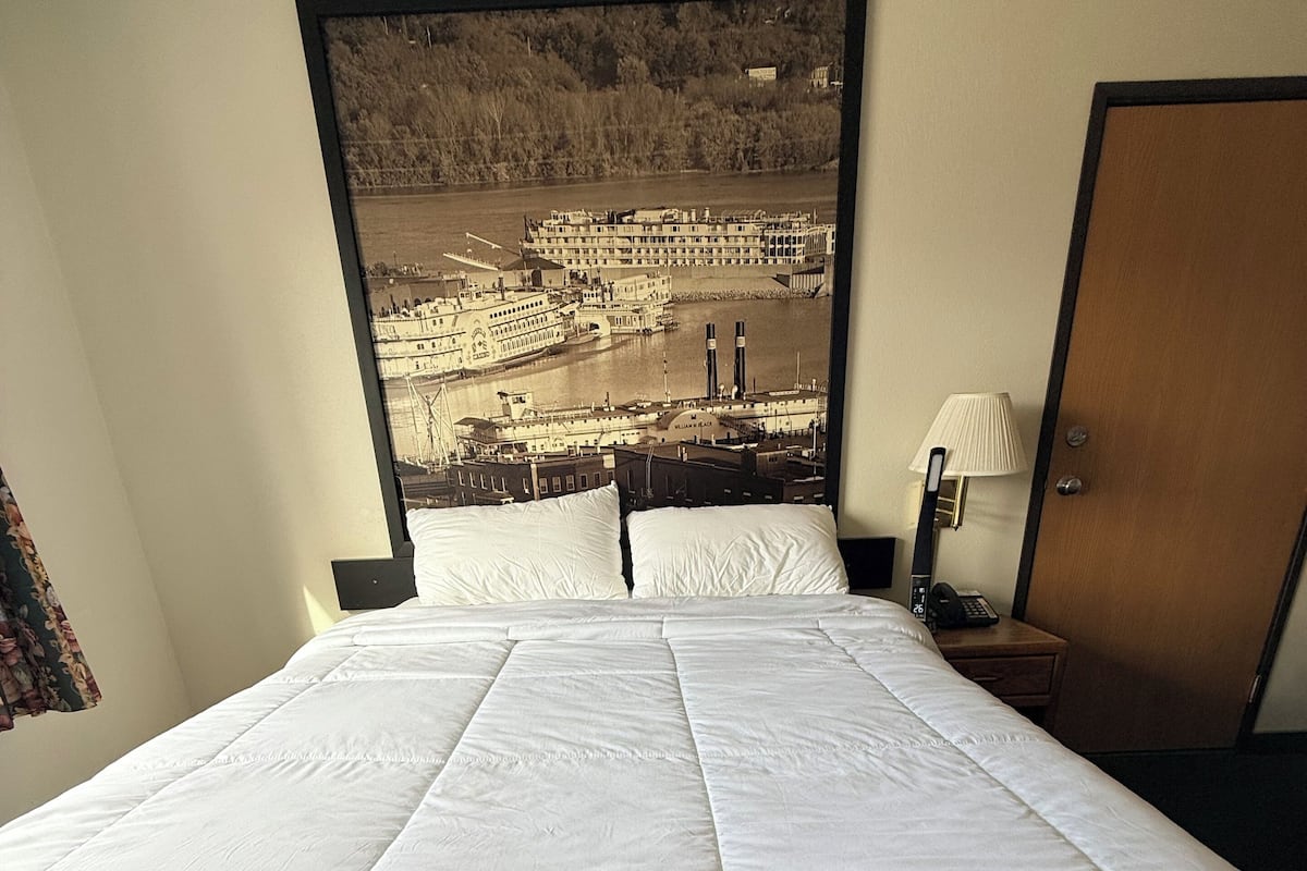 Et hotelværelse med en stor seng, en sengegavl med et bybillede, et natbord med en lampe og en dør.