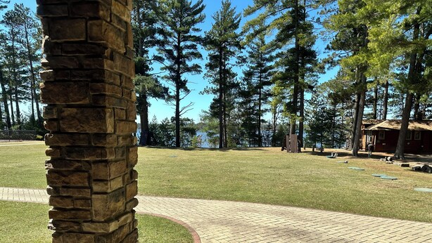A paved pathway leading to a grassy area with a stone wall on the left, surrounded by tall trees and a body of water in the background. - Spirit of the Northwoods (eagle river)