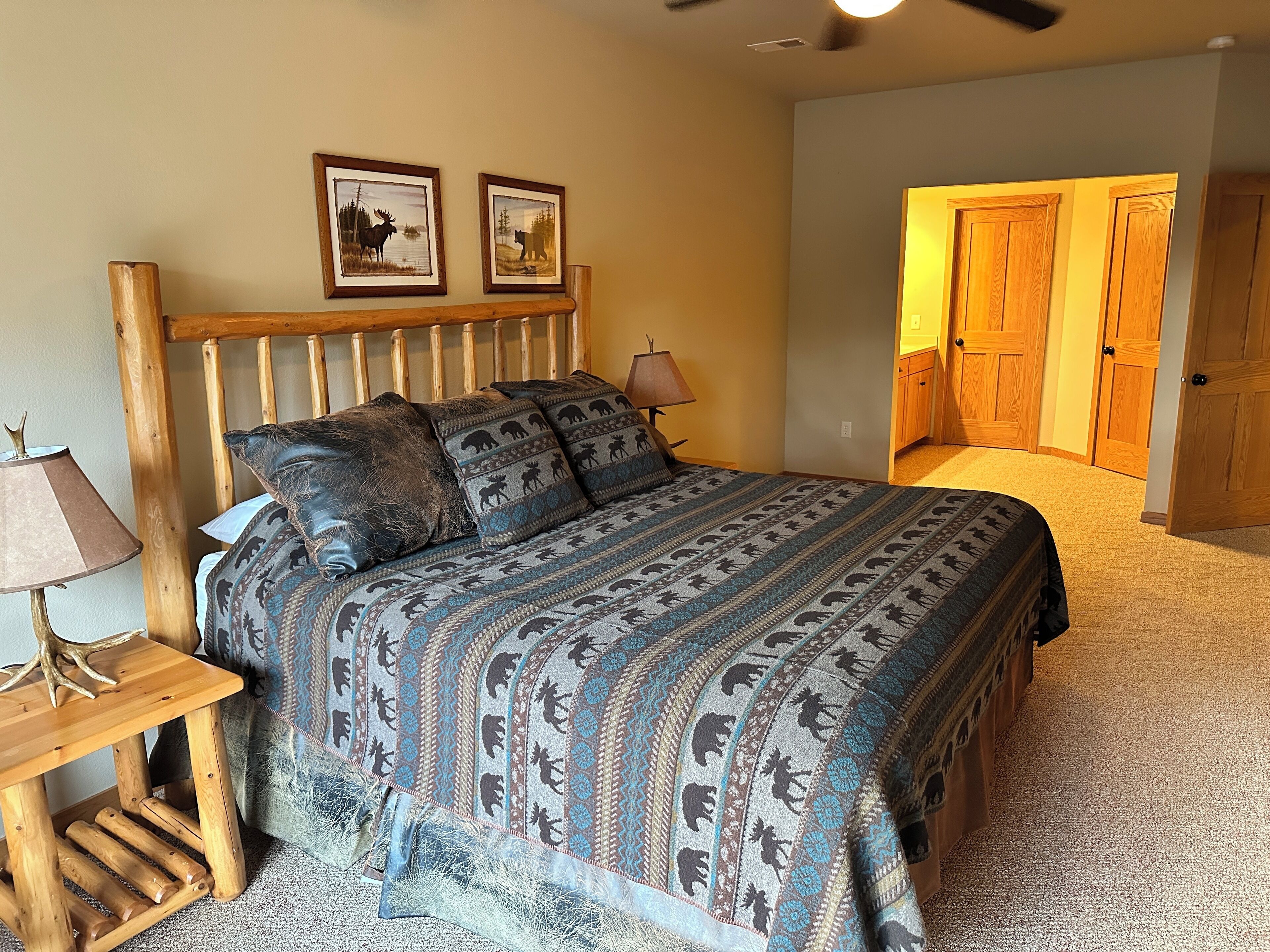 A bedroom with a wooden headboard, a patterned bedspread, two framed pictures on the wall, a bedside lamp, and a wooden nightstand.