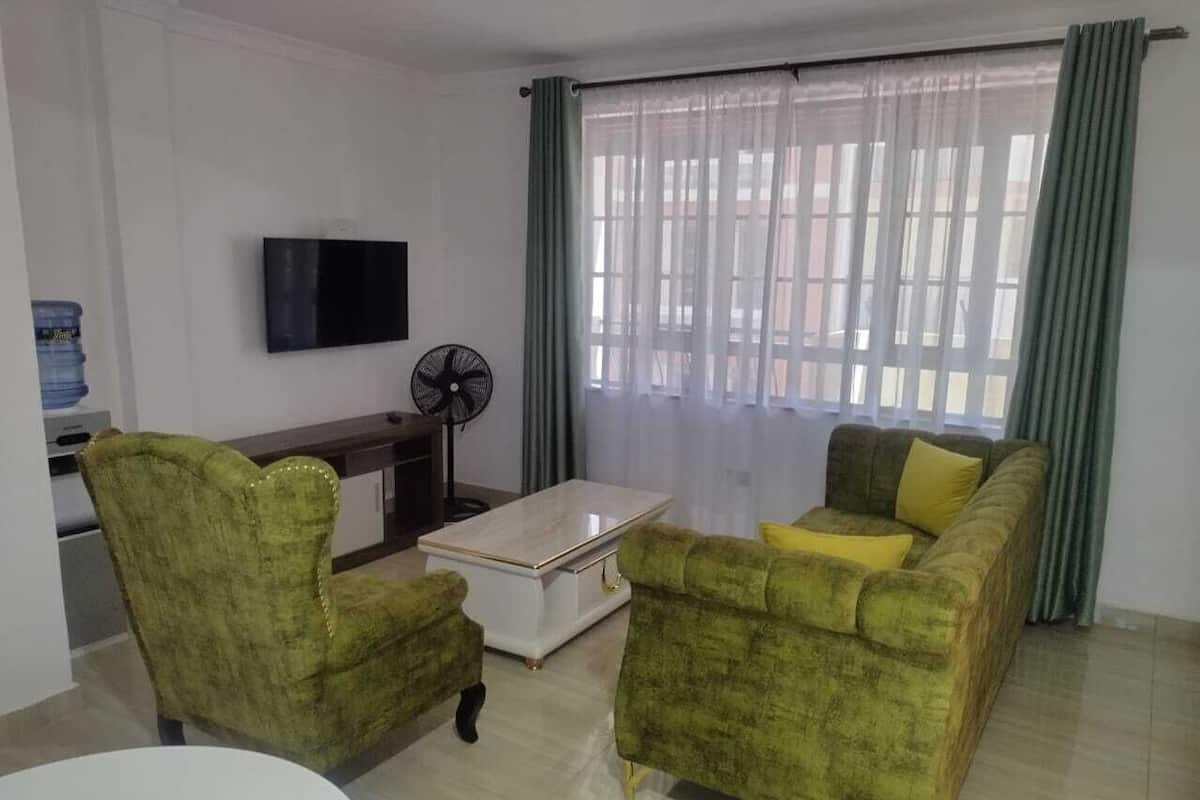 Una sala de estar con dos sillones verdes, una mesa de centro blanca, un televisor de pantalla plana montado en la pared, un ventilador de pie y una ventana con cortinas de gasa.