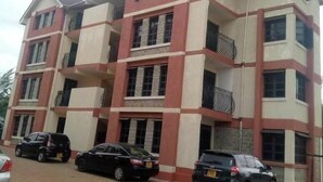 A multi-story residential building with a symmetrical facade, balconies, and a paved driveway with parked cars. - Nati Apartments Kisumu (Kisumu)
