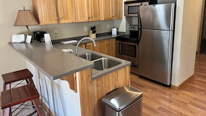 A compact kitchen with wooden cabinets, stainless steel appliances, a sink, and a small stool. - Spirit of the Northwoods (eagle river)