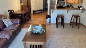 A modern kitchen with wooden cabinets, a stainless steel refrigerator, and a microwave. There's a dining area with a wooden table and chairs, and a leather sofa with a decorative bowl on it. - CONDO 107 by Eagle Waters Resort (eagle river)