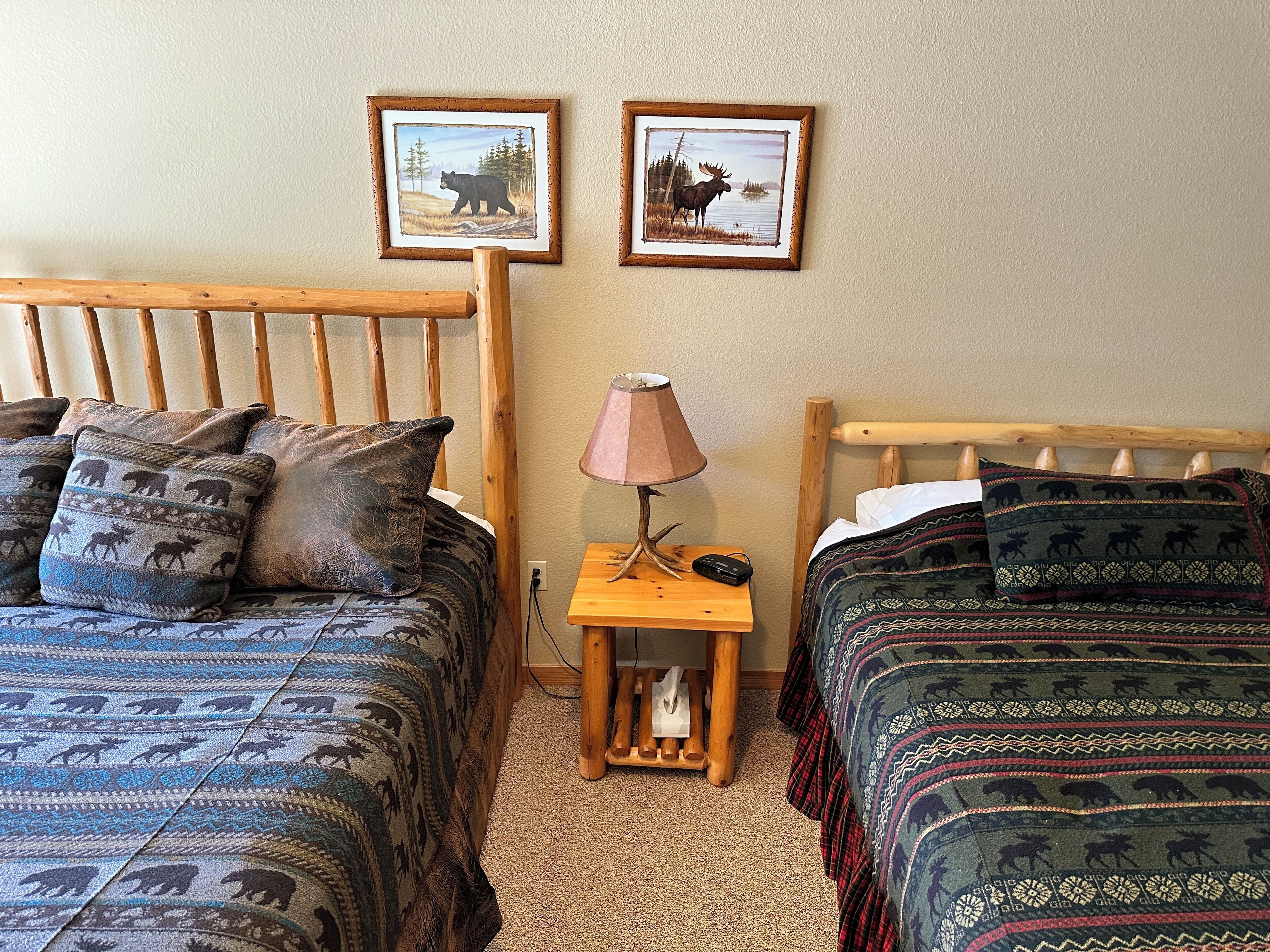 A room with two beds, each with patterned bedding, a wooden headboard, a bedside table with a lamp, and two framed pictures on the wall.