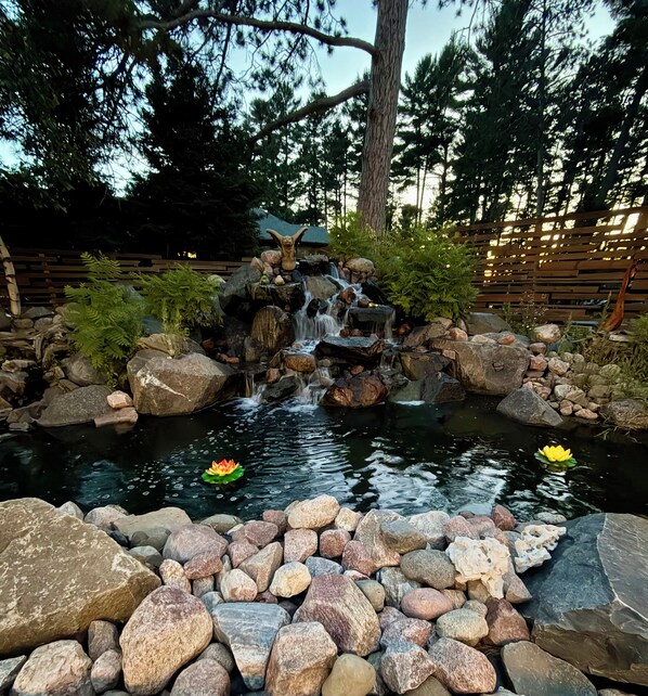 A tranquil garden with a small waterfall, a pond with lotus flowers, and a wooden fence.
