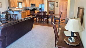 A modern kitchen with wooden cabinets, a dining area with a table and chairs, and a living room with a leather couch and a lamp. - CONDO 201 by Eagle Waters Resort (eagle river)