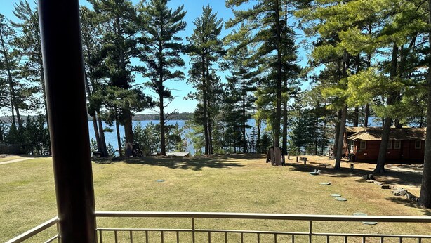 A view from a balcony overlooking a grassy area with trees and a lake. There is a wooden cabin and a metal railing in the foreground. - CONDO 201 by Eagle Waters Resort (eagle river)