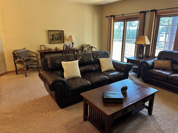 A living room with a dark leather sofa, a wooden coffee table, and a chair. There is a window with curtains, a lamp, and a framed picture on the wall.