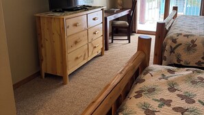 Une chambre avec un lit en bois, un téléviseur posé sur une commode en bois, un bureau avec une lampe et une fenêtre donnant sur l’extérieur.