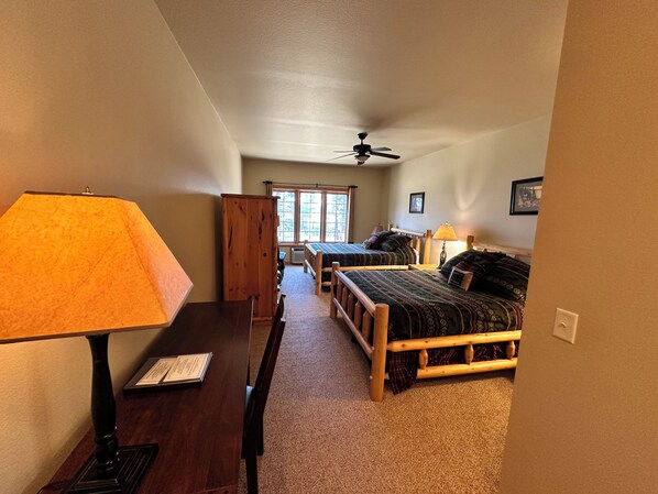 A bedroom with two beds, a wooden wardrobe, a desk with a lamp, and a ceiling fan.