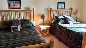 Una habitación con dos camas de madera, cada una con ropa de cama y almohadas estampadas. Hay dos cuadros enmarcados en la pared, una mesita de noche de madera con una lámpara, y un suelo alfombrado.