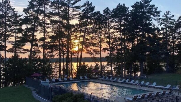 A pool area with lounge chairs and a view of the sunset behind trees. - Condo 206 W/ Garage by Eagle Waters Resort (eagle river)